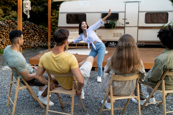 Joyful Asian lady playing word guessing game with diverse friends near camper van outdoors in autumn