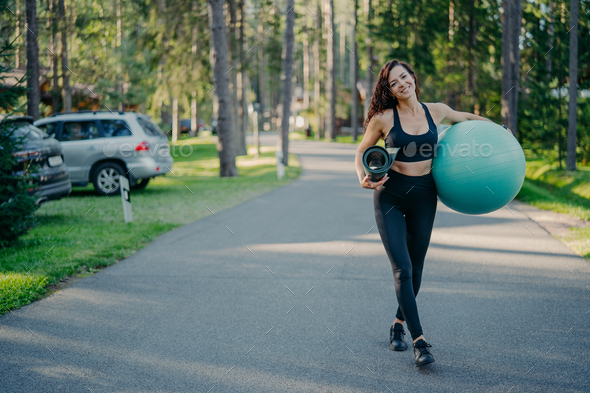 Active slim woman carries fitness ball and rolled up karemat, dressed in leggings