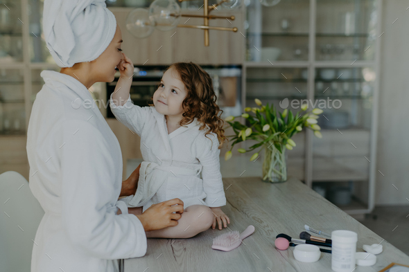 Girl touches nose of her mother dressed in white soft dressing gown sits on table with cosmetic