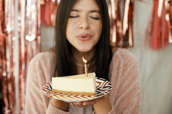 Piece of modern birthday cake with candle in hand of happy woman and pink tassel garland in room