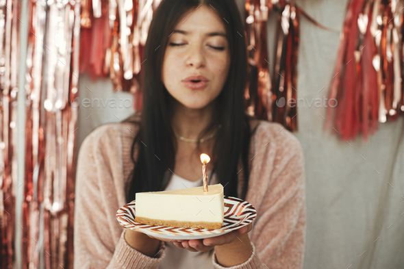 Make a wish. Stylish happy woman blowing burning candle on piece of birthday cake at tassel garland