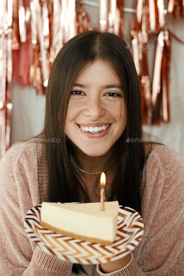 Stylish happy woman holding piece of birthday cake with candle at pink tassel garland in room