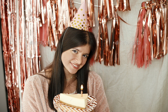 Stylish happy woman in party hat holding piece of birthday cake with burning candle at pink garland