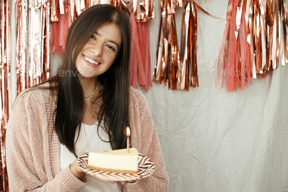 Stylish happy woman holding piece of birthday cake with candle at pink tassel garland in room