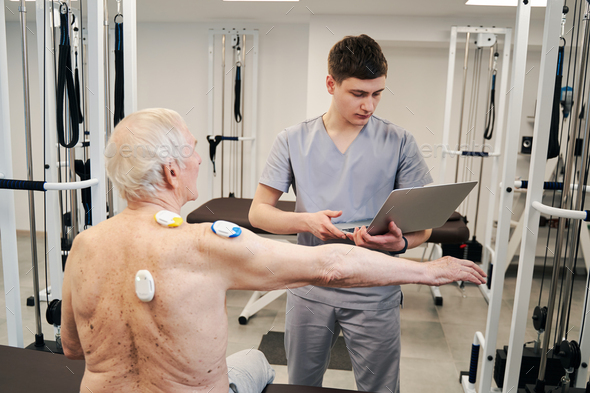 Physical therapist with laptop conducting back muscles diagnostics for retiree