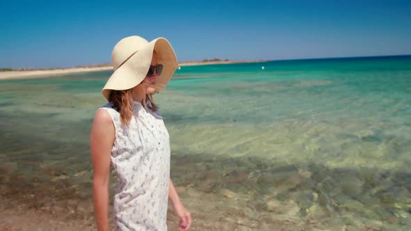 Free Woman in Hat and Sunglasses Walks Along Sea Shore on Sand Beach in Summer