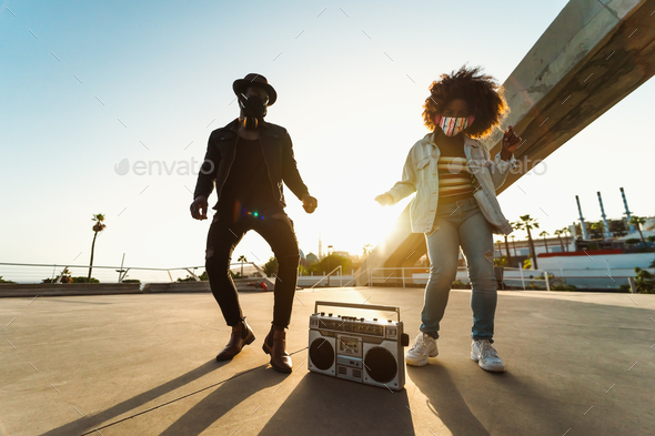 Young Afro friends wearing face mask dancing while listening to music with headphones and boombox