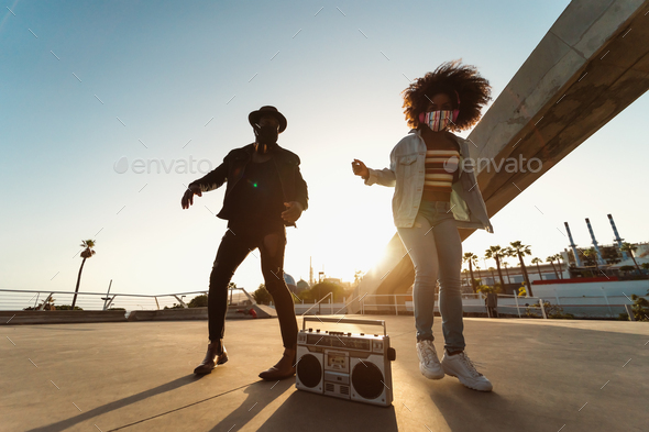 Young Afro friends wearing face mask dancing while listening to music with headphones and boombox