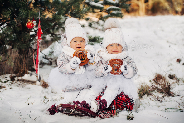 Two little twin girls in white suits sit and eat big buns in the winter outside