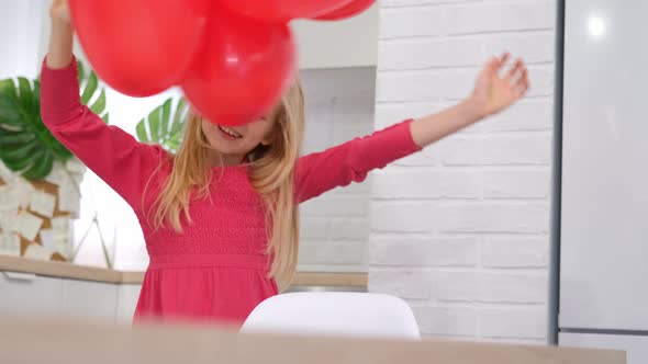 Little Blonde Girl Dancing with Red Heart Shape Balloons at Home alt