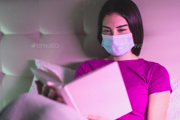 Young woman wearing surgical mask reading book in bed