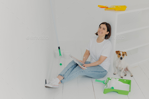 Woman in casual wear, sits on floor near white drawer, holds color samples, her dog poses near