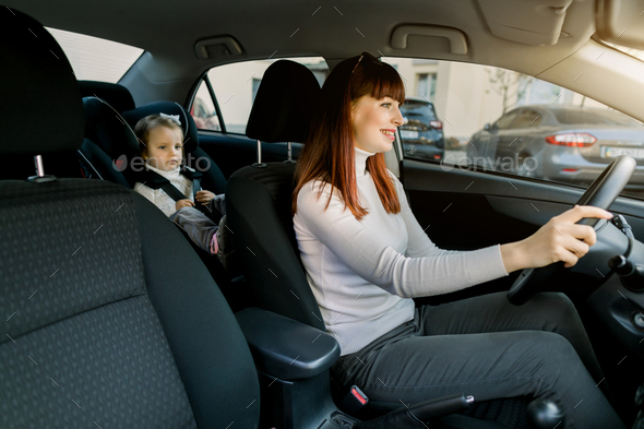 Pretty Caucasian woman, young mother driving a car, having her little baby girl in a child seat