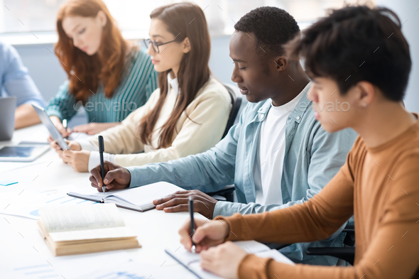 Diverse Students Sitting At Desk In Line Taking Notes