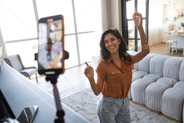 Cheerful female influencer shooting dance video for social media on mobile phone, having fun at home