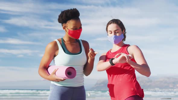Two diverse female friends wearing face masks holding yoga mats at the beach alt