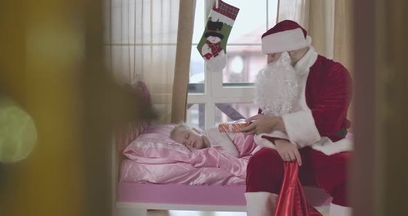 Santa Claus Taking Out Christmas Present and Putting It on Pink Pillow Where Cute Girl Is Sleeping alt