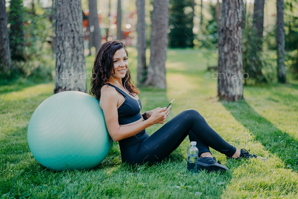 Woman sits back to big fitness ball, uses mobile phone for listening music,  poses on green grass Stock Photo by StudioVK