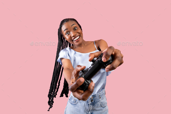 Modern entertainments concept. Excited young black lady with joystick playing video game on pink