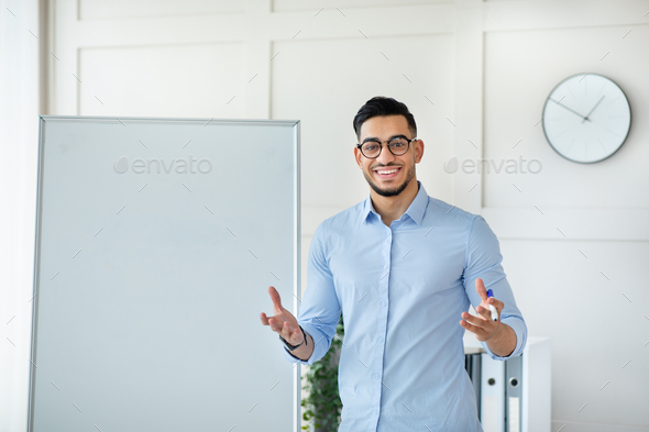 Joyful young Arab male teacher giving online private lesson from home, standing near empty