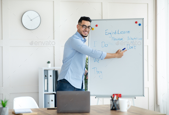 Young Arab male teacher pointing at blackboard with English grammar rules, giving online lesson to