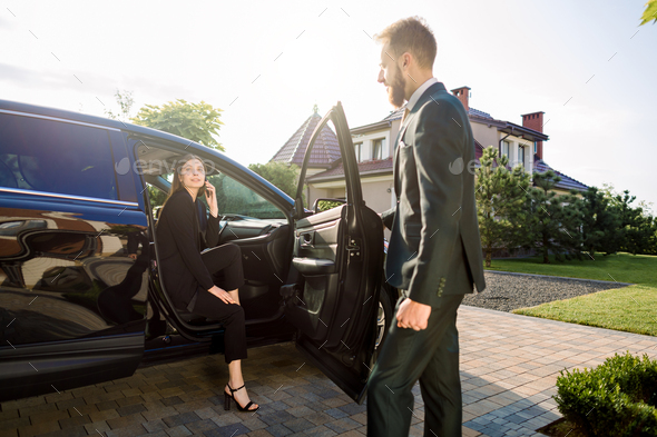 Young man personal driver waiting for his female boss, pretty young business lady in black suit, on