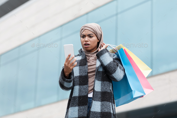 Surprised excited black lady with open mouth in hijab and coat with bags with purchase looks at