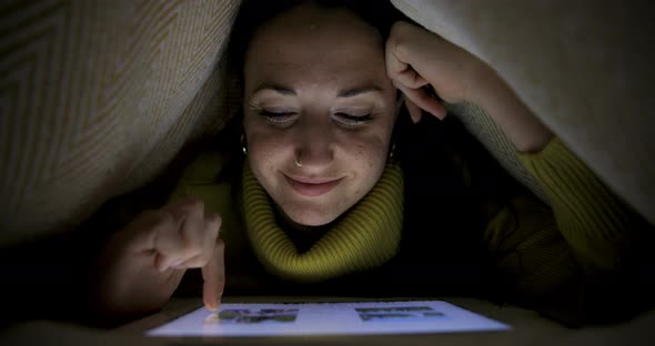 Woman on the bed under the blanket watching photos on digital tablet and smil