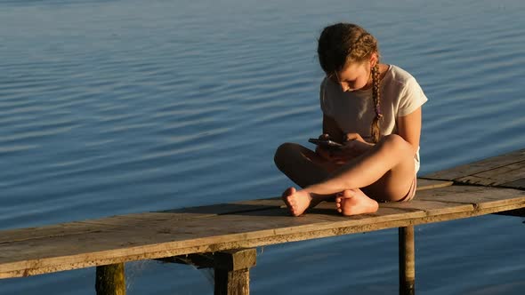 A little girl with a smartphone sits on a wooden pier on the lake during sunset. 
