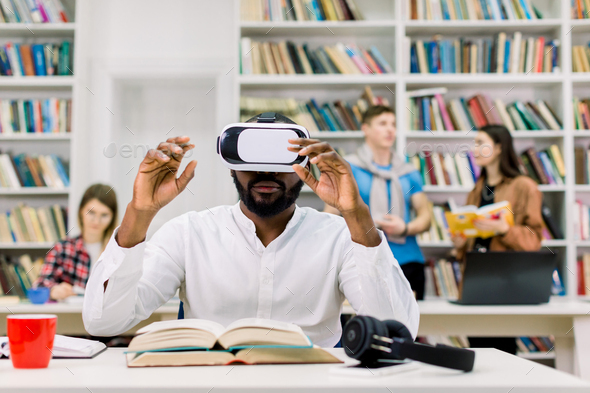 Male African student using VR goggles in public library and working with virtual textbook for