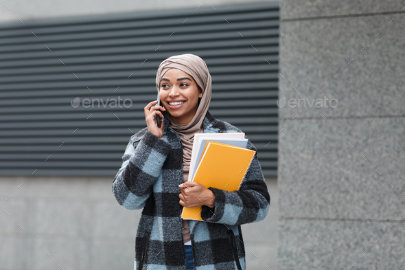 Smiling pretty muslim black lady student in hijab and coat with books, calls on phone in autumn