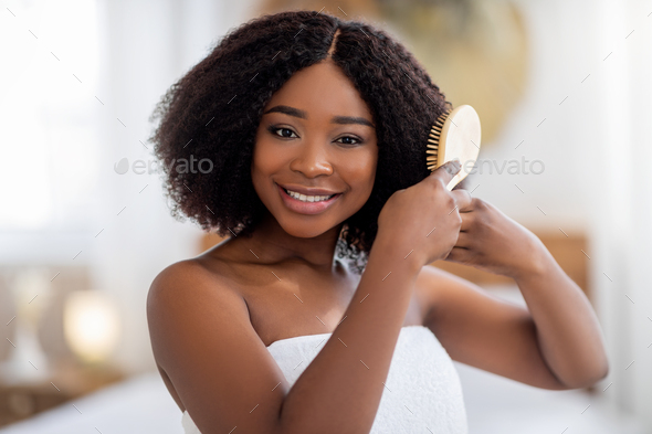 Portrait of beautiful young Afro lady brushing her dark hair, smiling at camera indoors