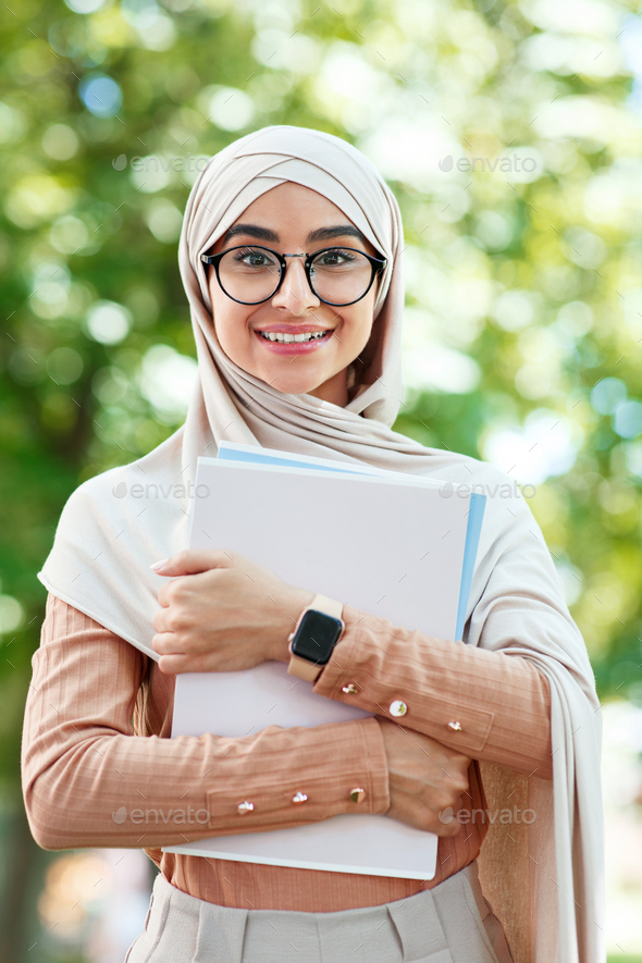 Cheerful glad cute young arab muslim female student in hijab and glasses holds books, ready for