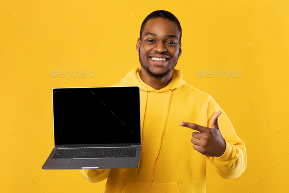 Black Guy Showing Laptop Computer Blank Screen Over Yellow Background