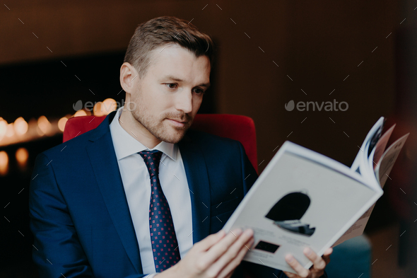 Concentrated young boss has stubble, being well dressed, reads interesting journal
