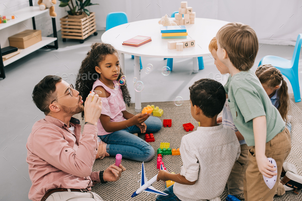 teacher with soap bubbles and multicultural preschoolers sitting on floor with colorful bricks in