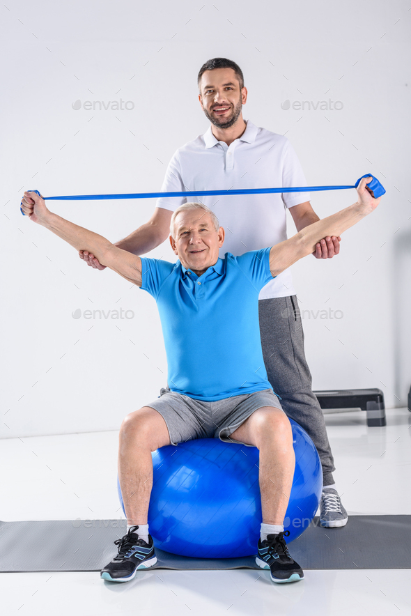 rehabilitation therapist assisting smiling senior man exercising with rubber tape on fitness ball
