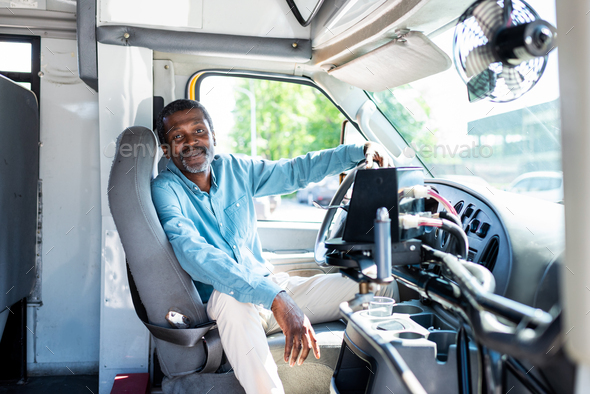smiling mature african american bus driver looking at camera while sitting inside bus