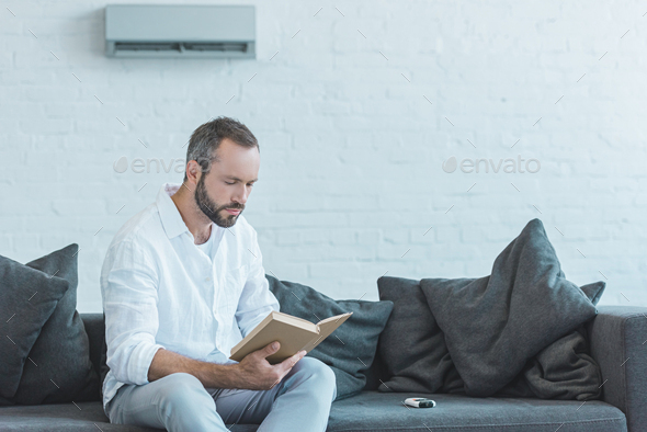 bearded man reading book on sofa, with air conditioner on wall