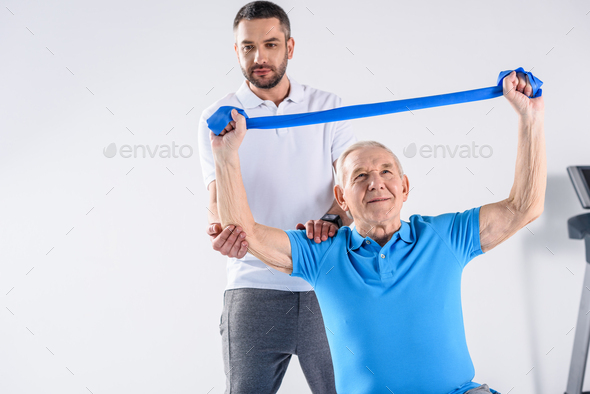 portrait of rehabilitation therapist assisting senior man exercising with rubber tape on grey