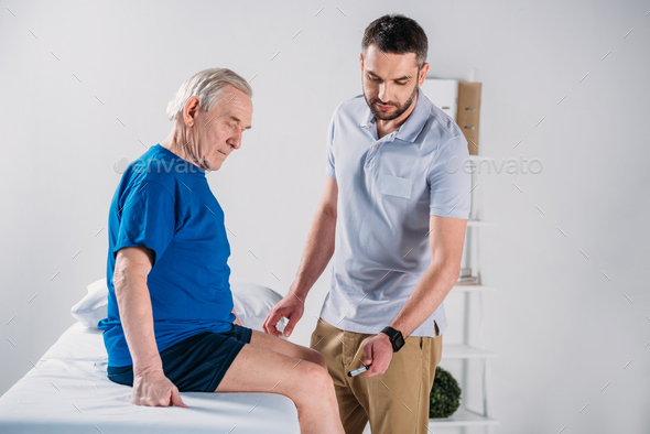 rehabilitation therapist with reflex hammer checking senior mans knee on massage table