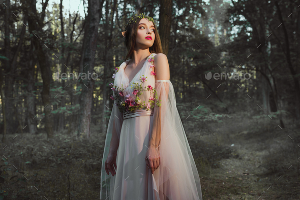 attractive mystic girl with elf ears posing in flower dress in forest