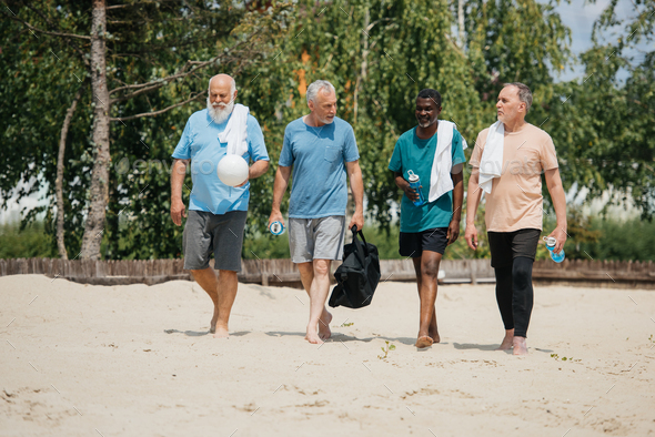 multiethnic elderly volleyball players with sportive water bottles walking on beach after game