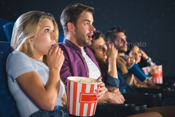 scared multiethnic friends with popcorn watching film together in movie theater