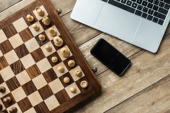Smartphone, laptop and chess board with chess pieces on wooden surface