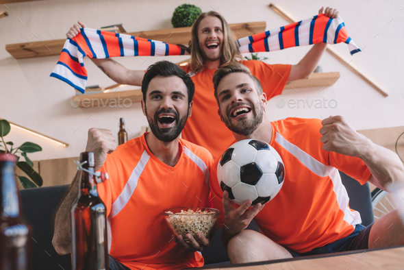 excited young male football fans in orange t-shirts with scarf, ball and popcorn celebrating and