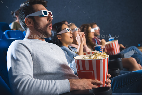 shocked multiethnic friends in 3d glasses with popcorn watching film together in movie theater
