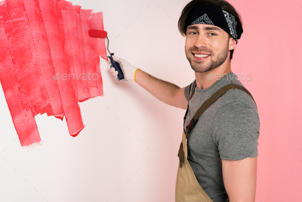 smiling man in working overall and headband painting wall in red by paint roller