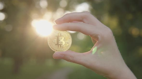 Man Shows a Cryptocurrency Symbol Bitcoin Coin on the Background of a Green Blurred Trees Outdoors