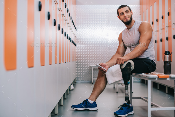 smiling young sportsman with artificial leg sitting on bench at gym changing room and using